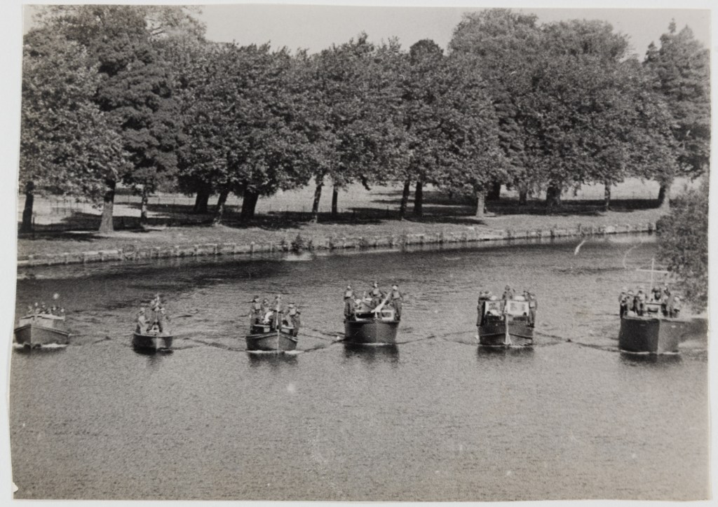 Black and white photograph of six small boats in a row on the Thames, carrying several passengers. In the background there is a row of trees at the water’s edge. 