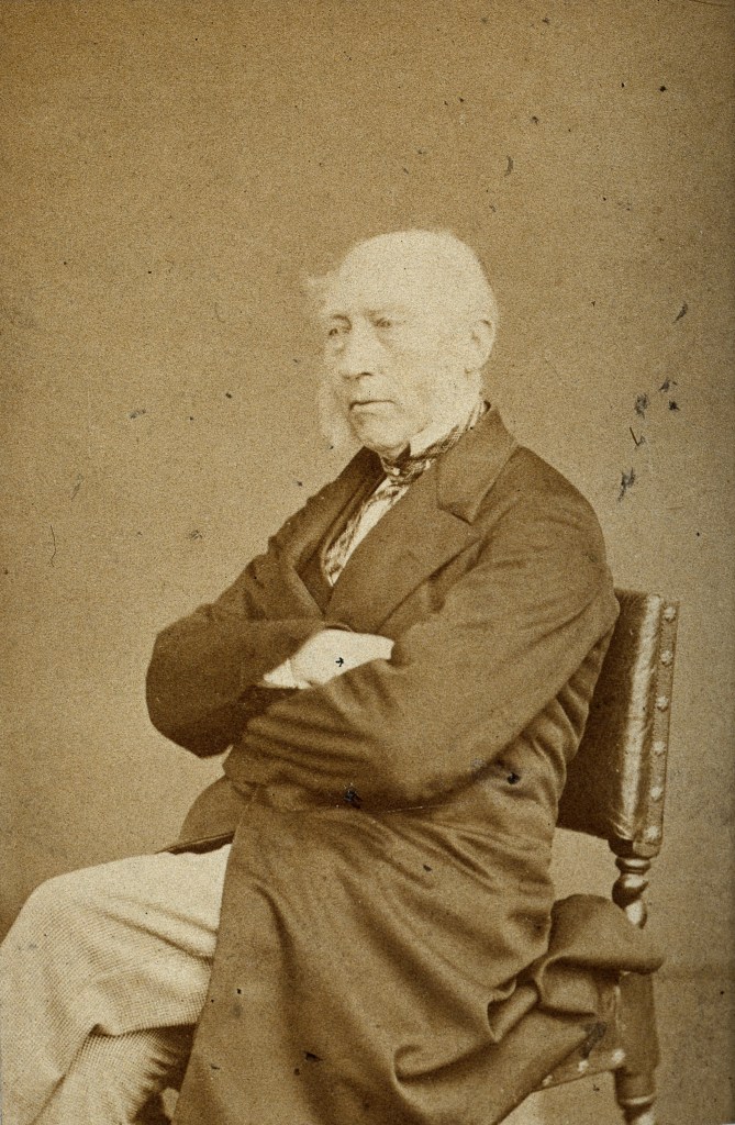 Sepia photograph of Professor John Phillips dressed in smart attire and sat on a chair with folded arms, looking away from the camera.