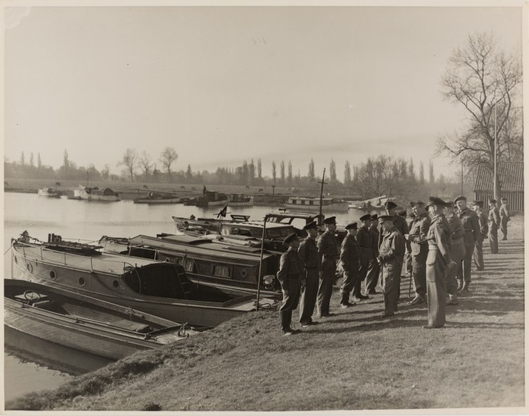 Black and white photograph of soldiers in uniform stood to attention in line before their superiors along the riverbank. Behind them is a row of small boats moored on the riverside.