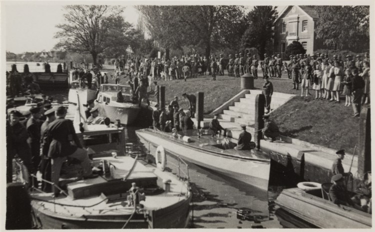Black and white photograph of boats by the riverside with soldiers on board. On the riverbank is a set of stone stairs leading to a large building. There is a crowd of civilians and soldiers watching the boats.