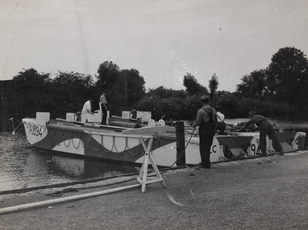 Black and white photograph of a moored landing craft. Two men are stood on the riverbank, holding rope and holding the side of the boat. Three men are aboard the boat.