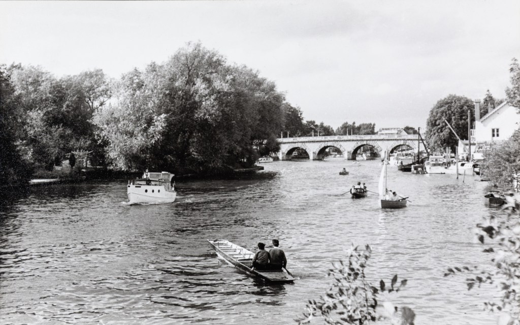 Black and white photograph of boats on the River Thames with an arched bridge in the background. In the foreground is a long wooden paddle boat with two boys sat side by side at the back. There is also a small motorised boat and other paddle boats on the river.