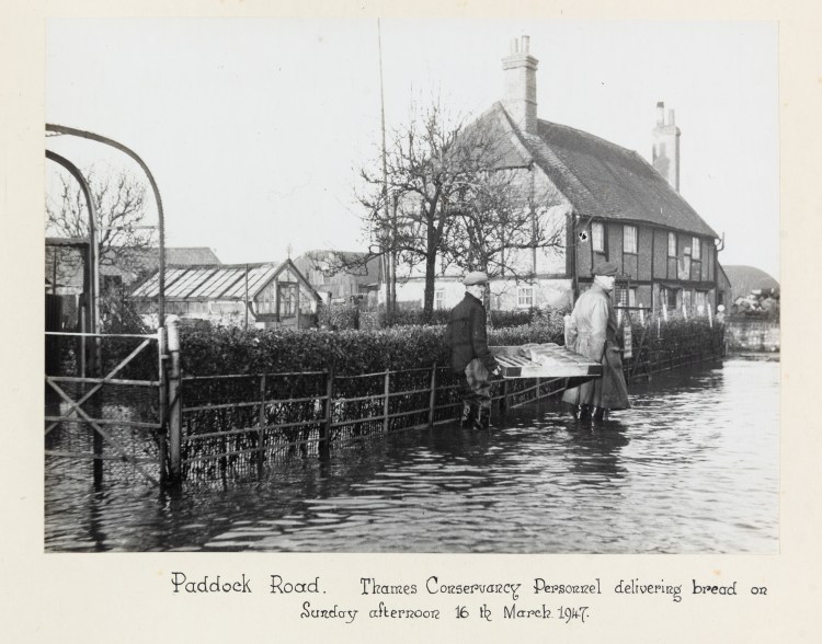 Black and white photograph of two men carrying a tray of bread through a flooded street with houses in the background.