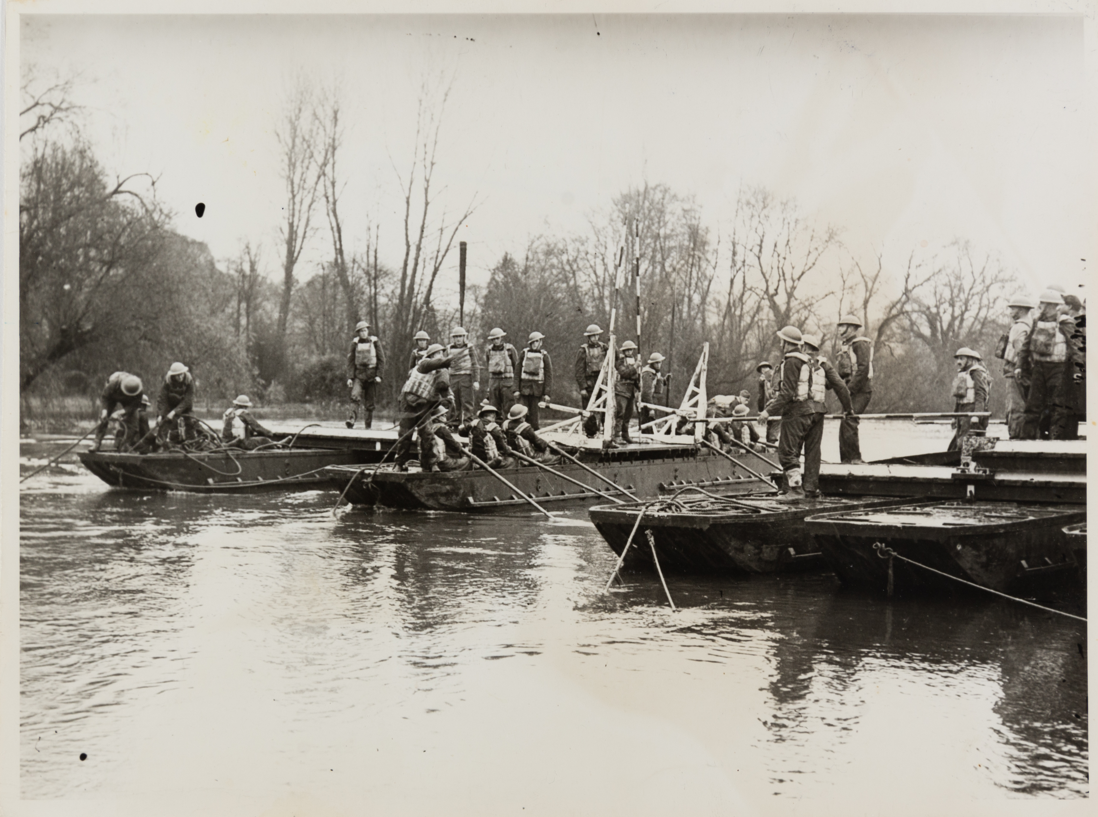 Black and white photograph of several soldiers standing and sitting on moored landing crafts in the Thames. Some are pulling rope or holding poles and others are sat down holding paddles.