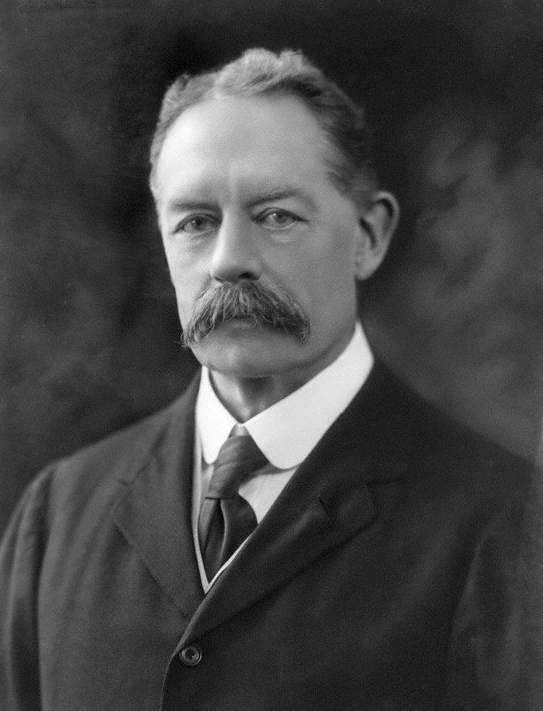 Headshot of a smartly dressed man in his mid 60s with a moustache and parted hair wearing a suit jacket, shirt and tie.