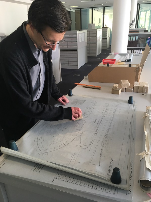 A man in his 30s cleaning a construction plan on a large table using a small smoke sponge. A pile of sponges, a large brush and a box are on the table next to him.