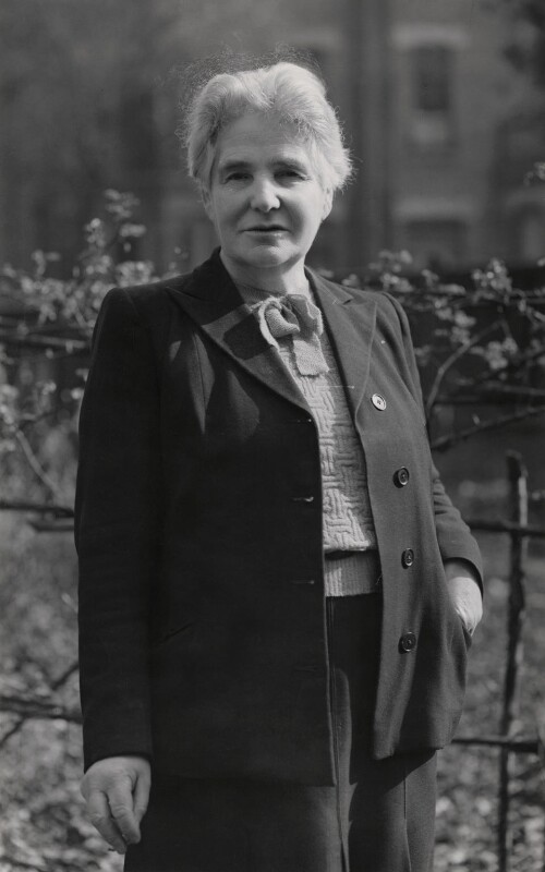 A woman in her late 50s with short white hear wearing a smart jacket, trousers and knitted sweater with a bow at the neck. 