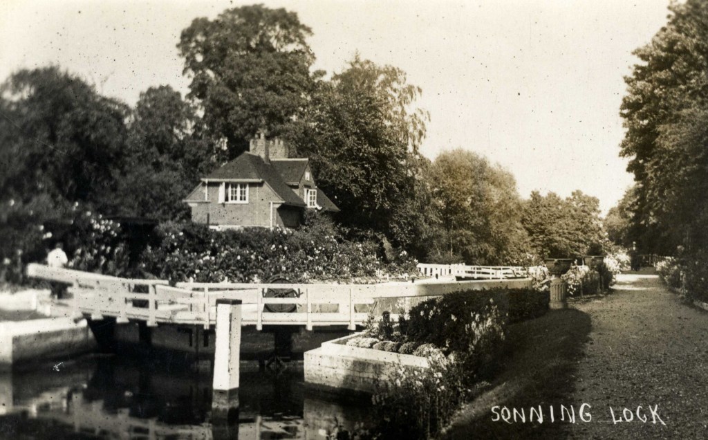 Black and white photograph of a brick house behind a lock bridge. Flowering shrubbery surrounds the house and bridge.